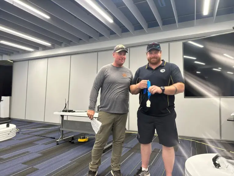 Inside a gray room, a man wearing a black shirt and shorts stands alongside a man in a gray shirt and pants