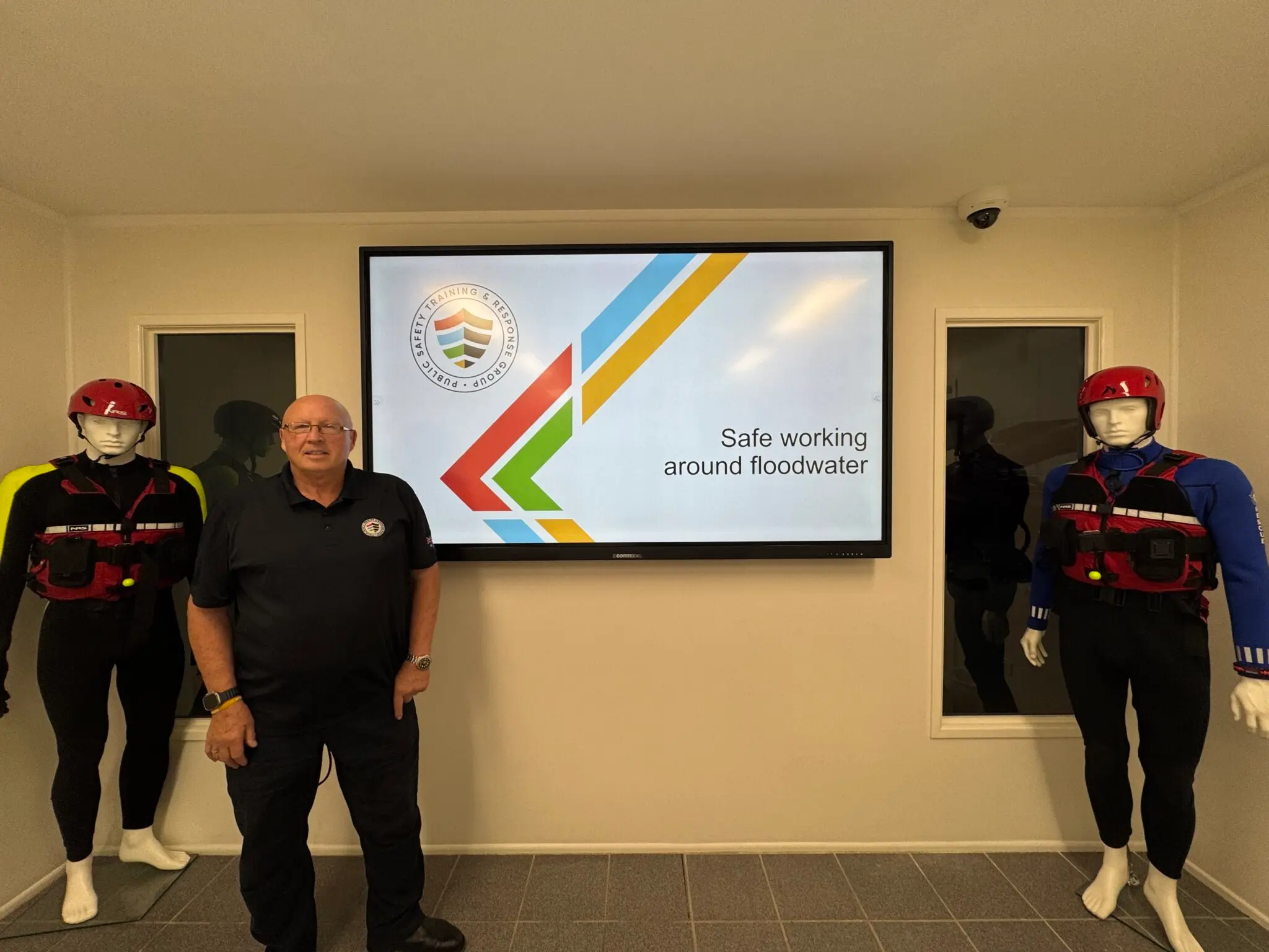 In a white room, a man dressed in black stands before a monitor, flanked by mannequins in red vests and red hats on either side