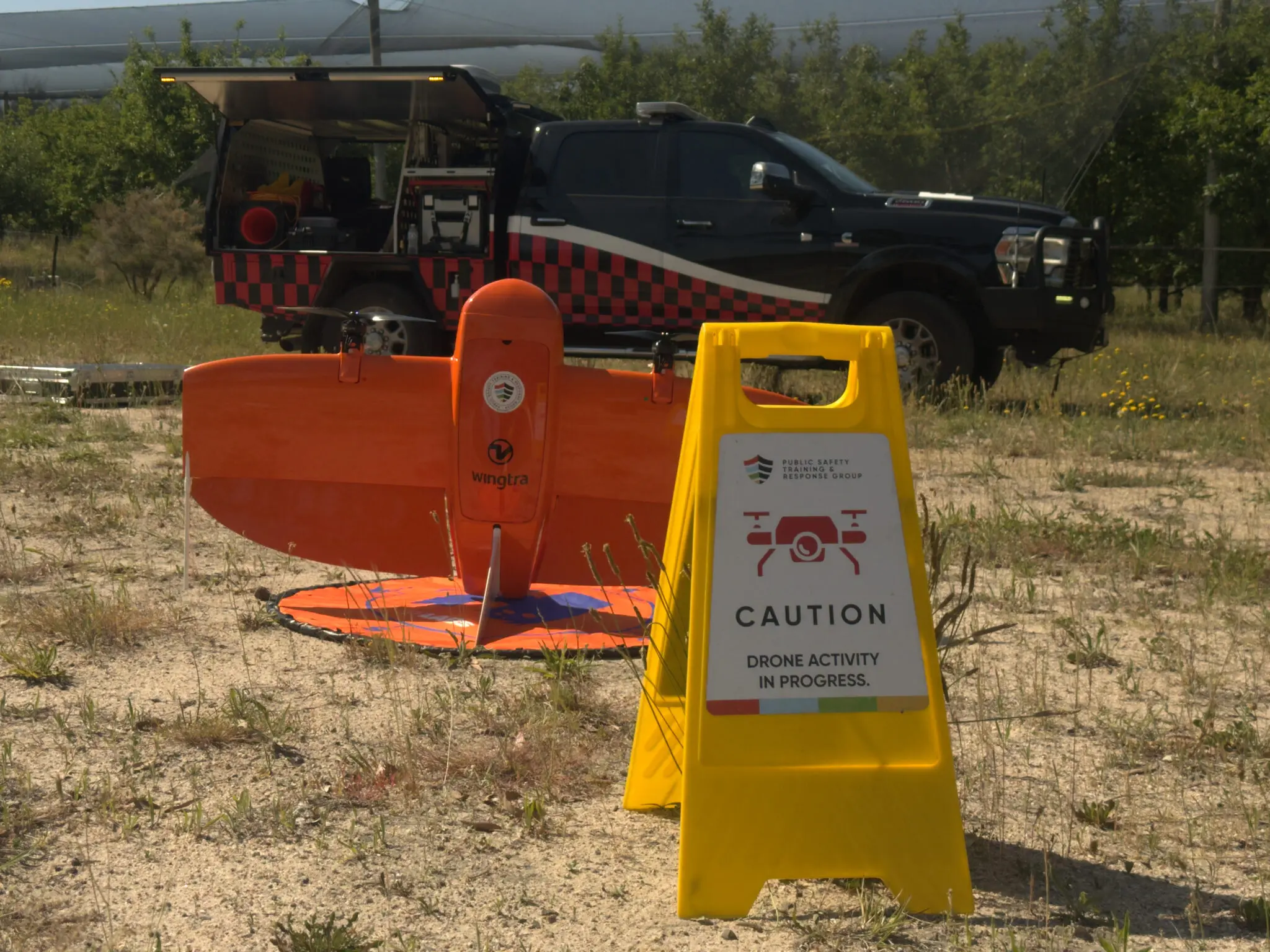 An orange airplane-shaped rocket is positioned on the ground before a black pickup truck with a red stripe, with a yellow cone placed in front
