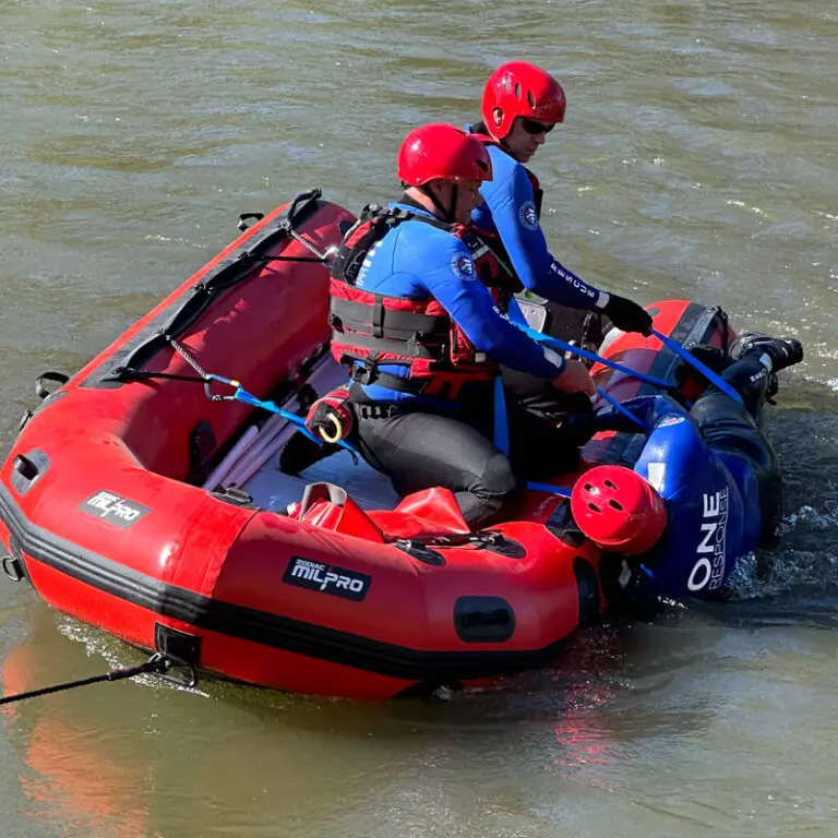 Two men in blue shirts, red vests, and red hats on a red inflatable boat are rescuing someone from the water