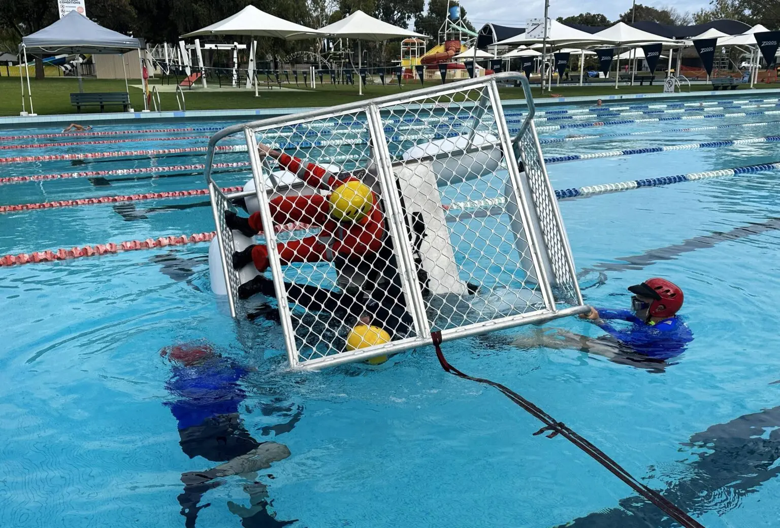 A man in an orange shirt is inside a cage in the middle of a pool, while a man in a blue shirt, red vest, and red hat is outside the cage