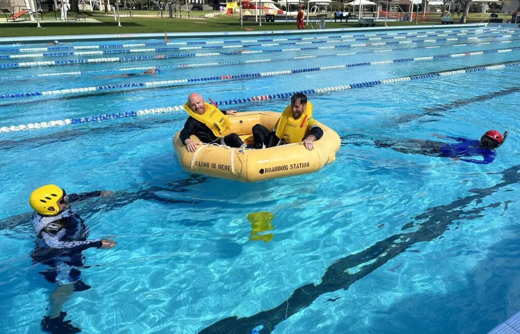 Two men wearing black shirts and life jackets are on a yellow inflatable boat in the center of a pool.