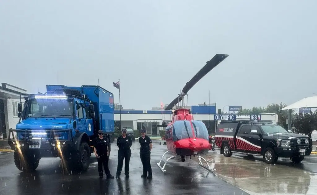 On the left is a blue truck, in the center a red and black helicopter, and on the right a black pickup, with three people wearing black shirts standing in front