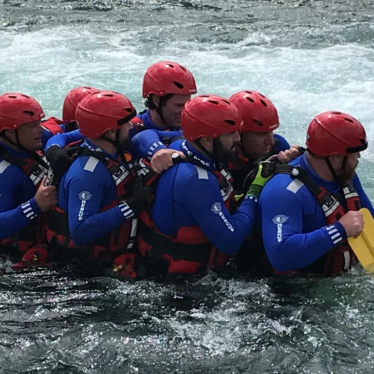 Seven people in blue shirts, red vests, and red hats are rowing a boat upstream against the strong current in a canal