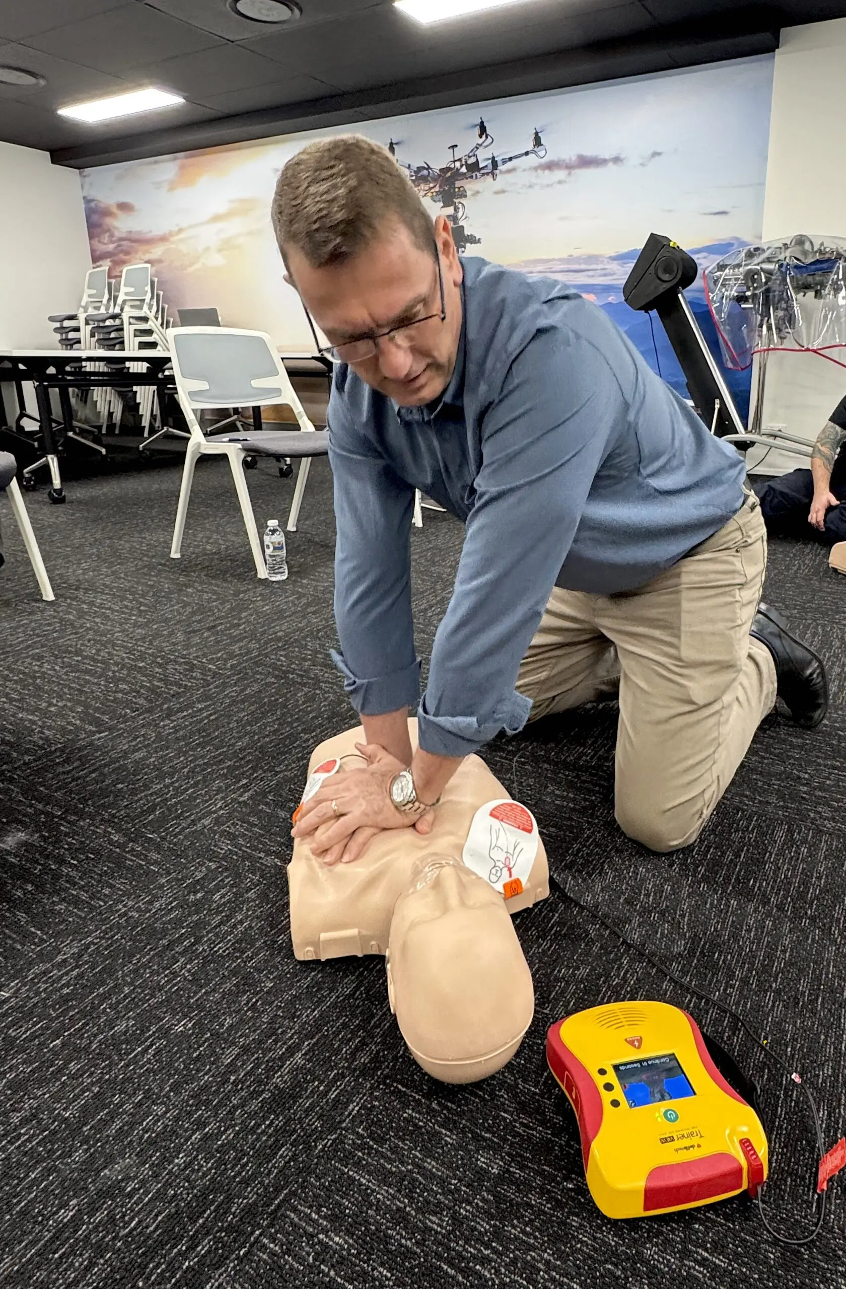 A man wearing a blue shirt and beige pants is practicing CPR on a male mannequin.