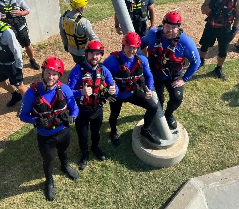 Four men in blue shirts, red vests, and red hats stand by a pole, surrounded by people wearing gray shirts