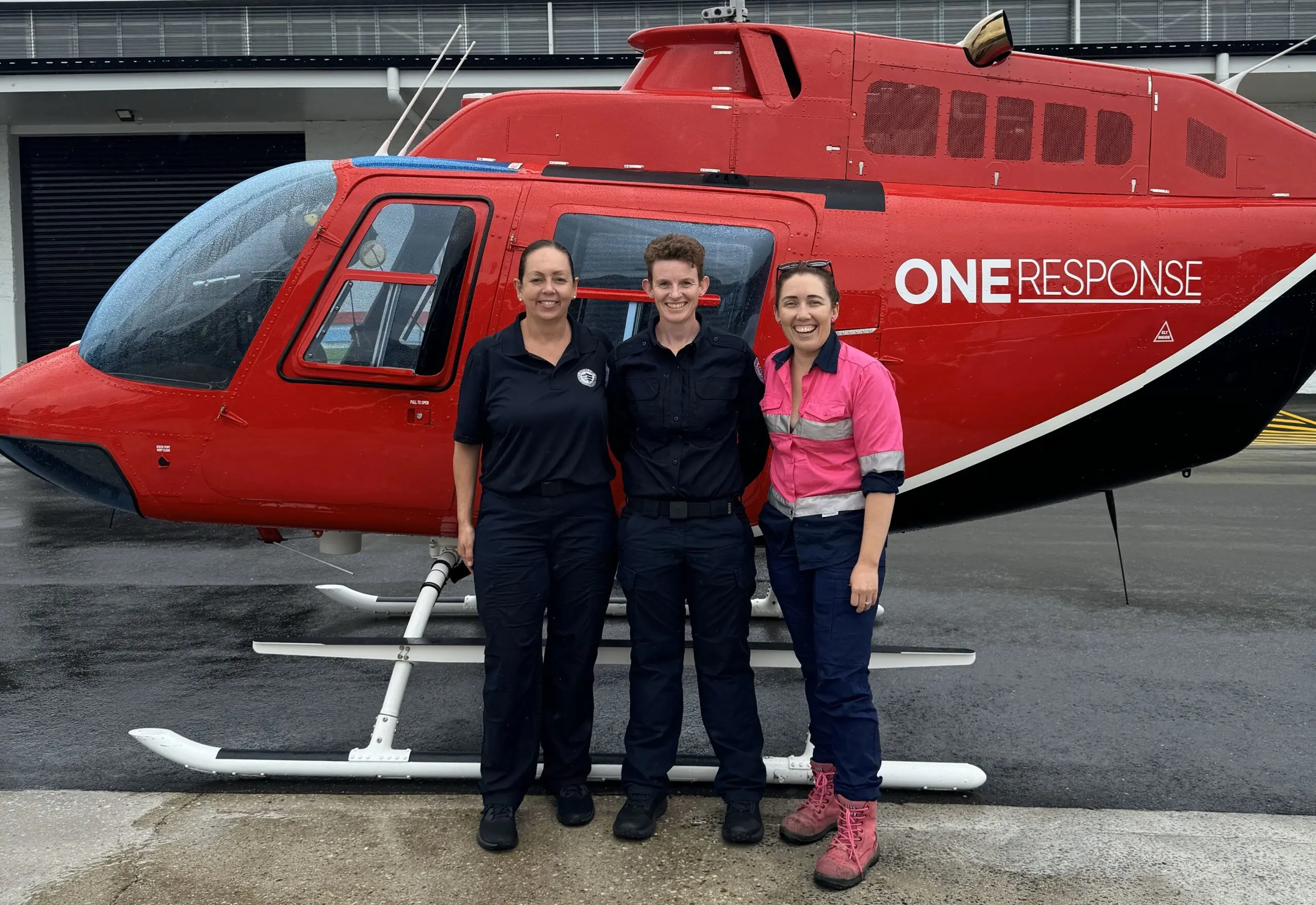 Three women stand before a red and black helicopter